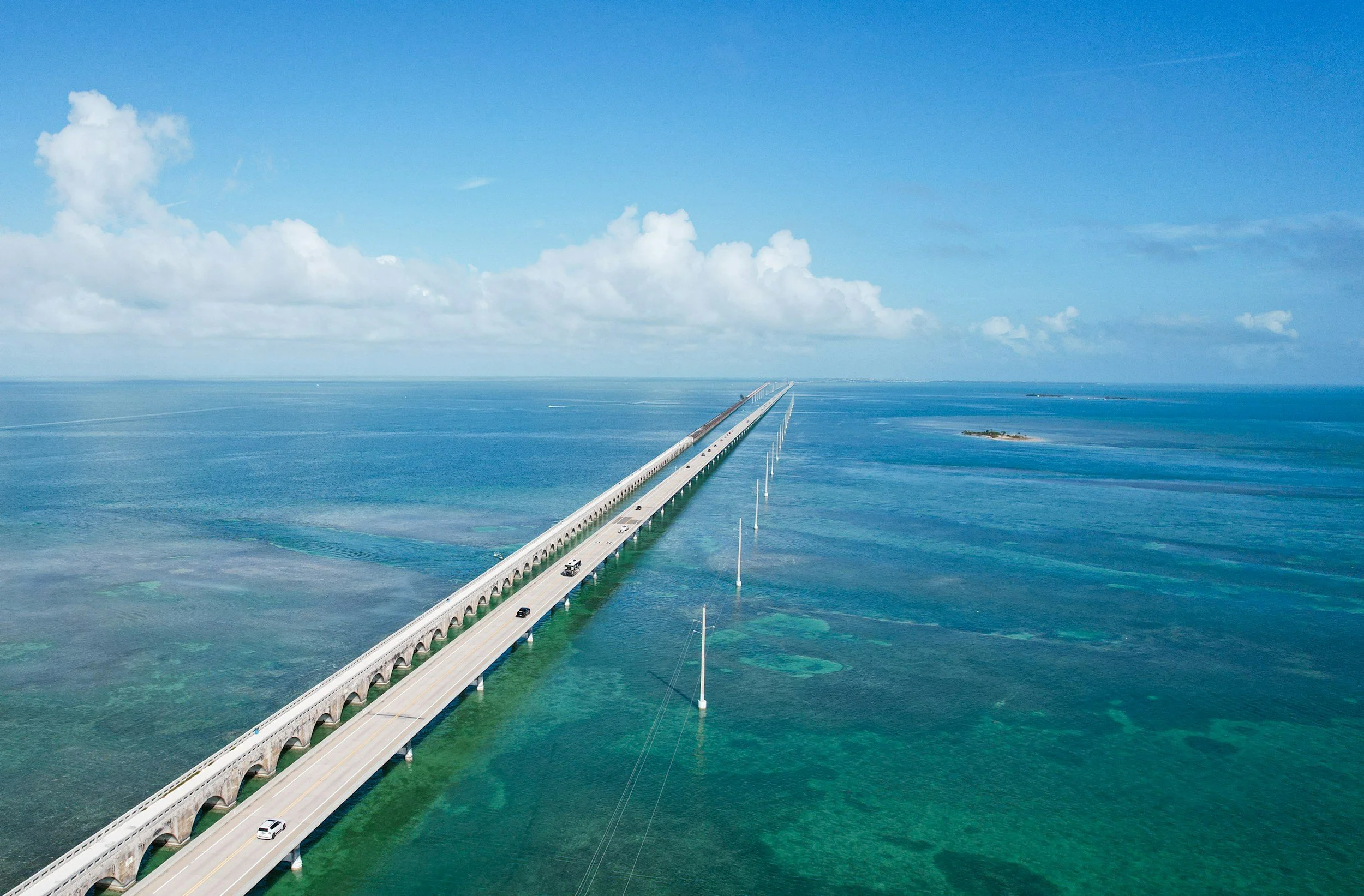 An aerial view of a long bridge extending over turquoise ocean waters, with a few cars driving on it under a partly cloudy blue sky.