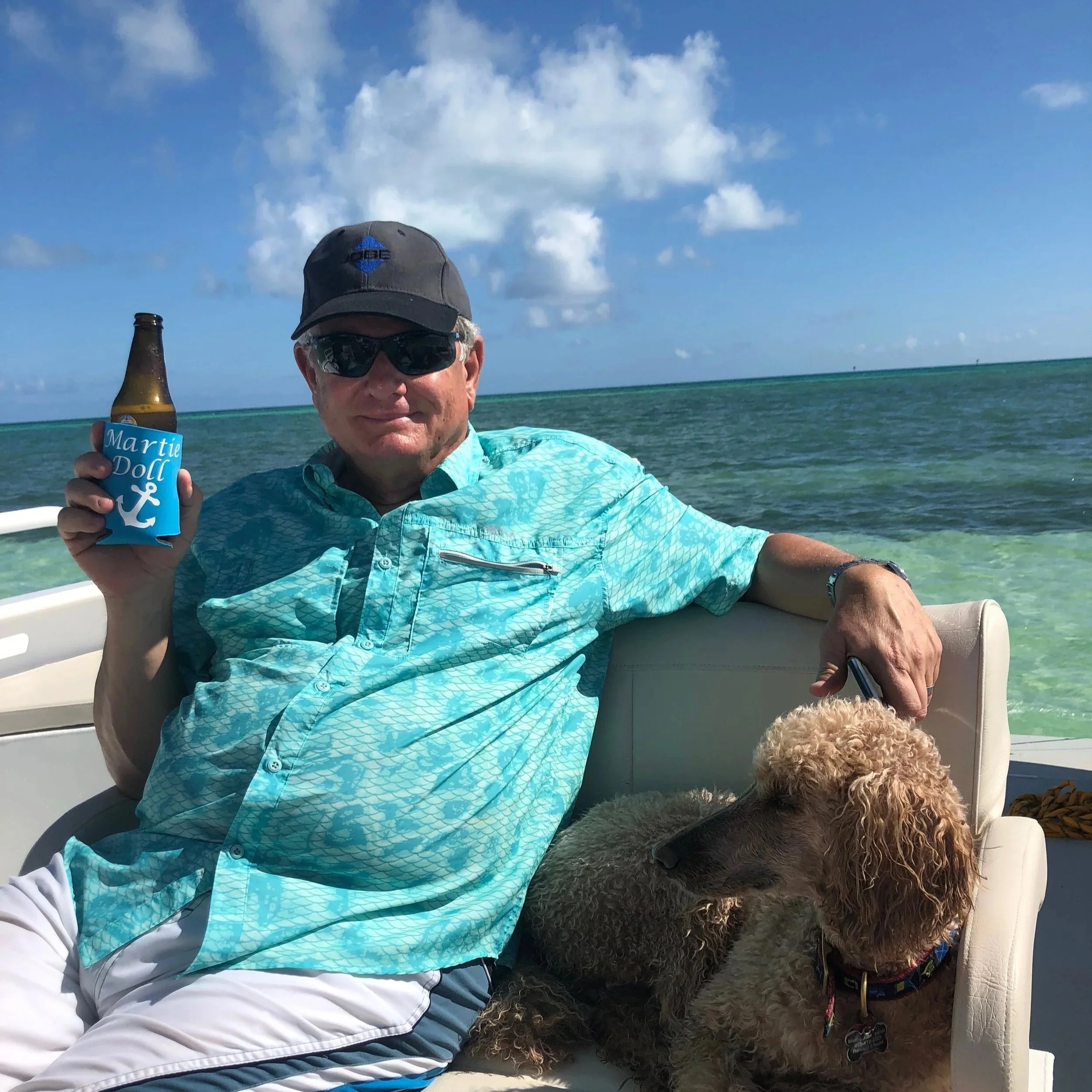 A man wearing sunglasses, a cap, and a turquoise shirt sitting on a boat with a poodle dog. He is holding a bottle with a blue koozie that says 'Martie Doll' and an anchor symbol. The background shows water and a partly cloudy sky.