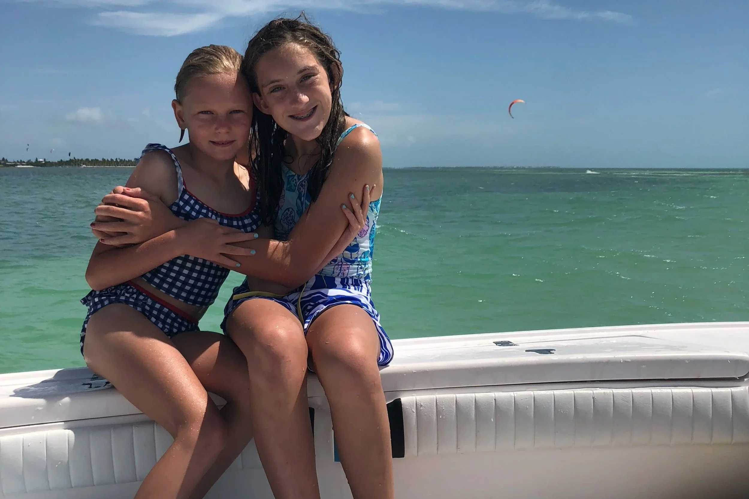 Two girls sitting on a boat by the ocean, hugging and smiling at the camera, with a kite surfer and blue sky in the background.