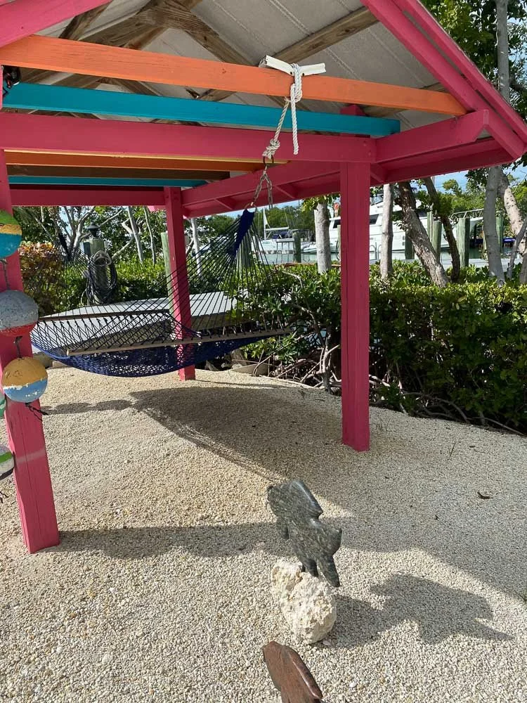 Colorful wooden structure with pink, orange, turquoise, and yellow beams, featuring a black hammock hanging underneath on sandy ground with a decorative frog sculpture and greenery in the background.