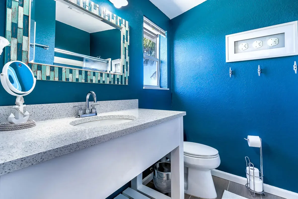 Bathroom with blue walls, white sink with speckled countertop, mirror with patterned frame, small window, toilet, toilet paper holder with roll, and wall-mounted clock with four circular dials.