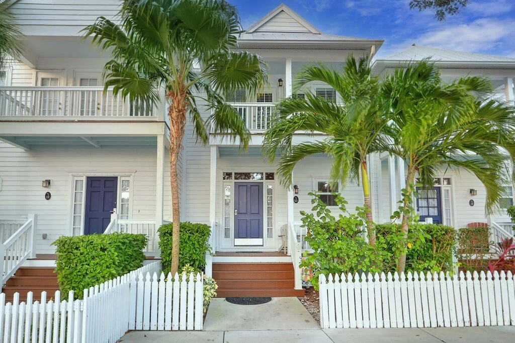 Front view of a white house with blue doors, a small front porch with stairs, green hedges, tall palm trees, and a white picket fence, under a partly cloudy sky.