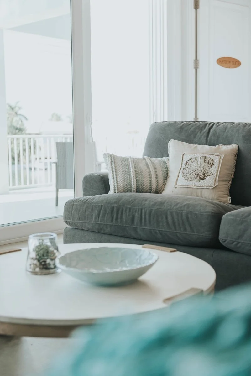 Living room with a gray couch with decorative pillows, a round coffee table with a bowl and a small plant, and large glass sliding doors leading to an outdoor balcony with a chair.