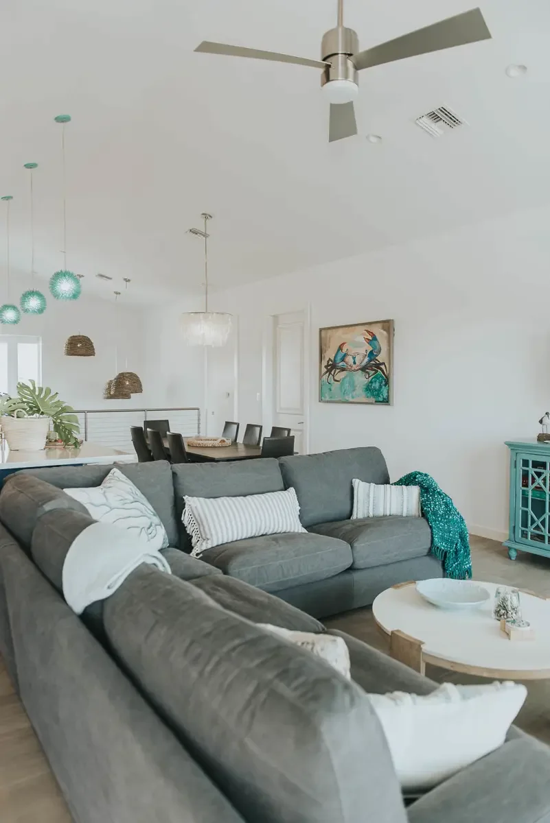 A living room with a gray sectional sofa, white and striped pillows, a round coffee table, a turquoise sideboard, a painting of two birds on a branch, a ceiling fan, and light fixtures hanging above the dining area.
