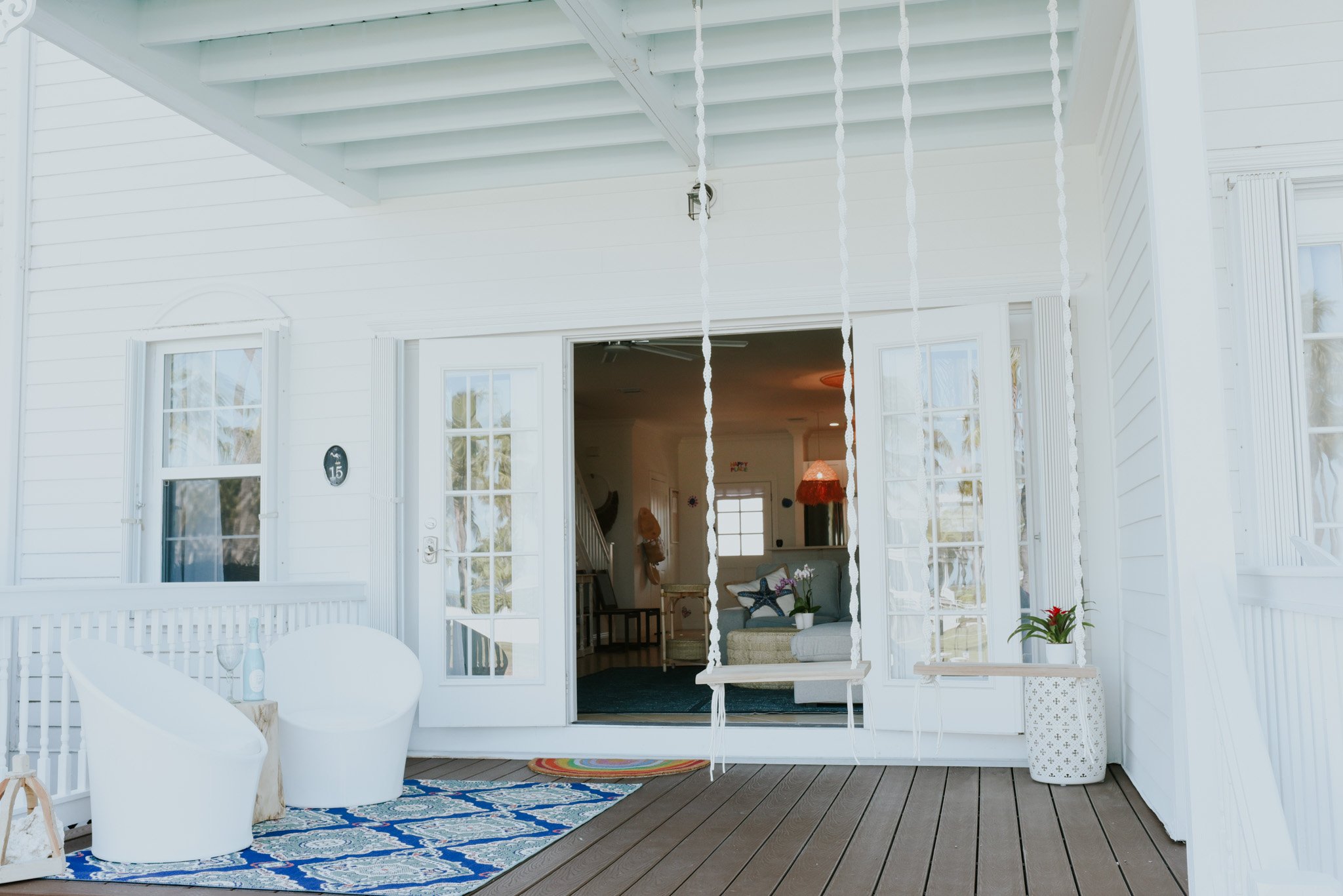 A white porch featuring two white chairs, a potted plant, and a colorful rug, with an open door revealing an interior living space with a sofa, hanging lamp, and decorative items.