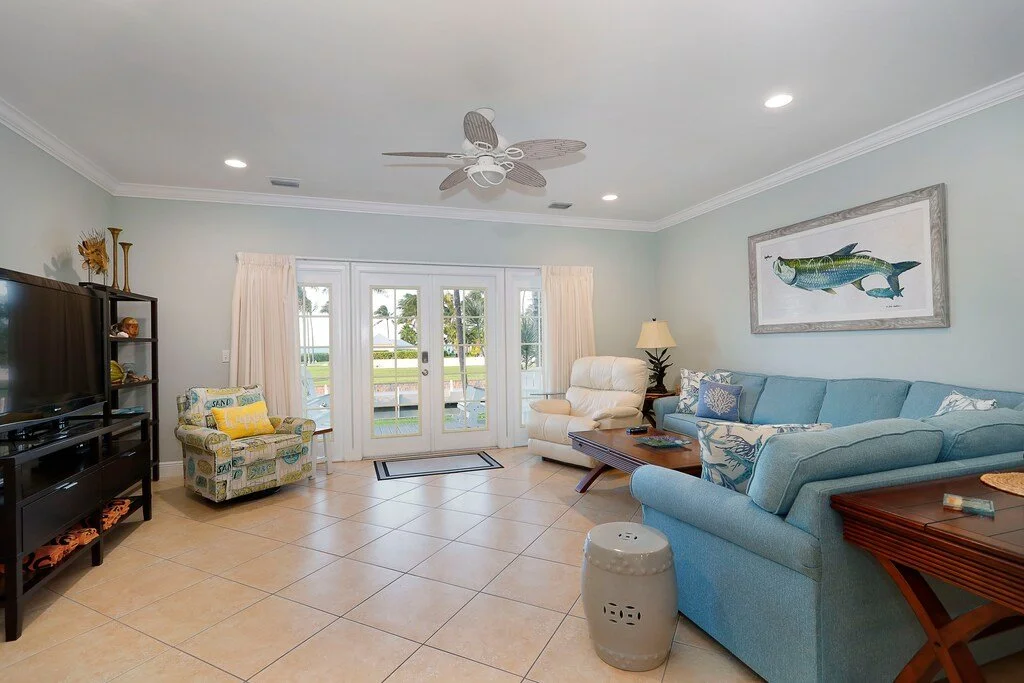 Living room with light green walls, large blue sectional sofa, white leather armchair, and a patterned armchair. French doors leading to a patio with outdoor furniture and a view of a canal. Fish artwork on the wall, ceiling fan, and various side tab