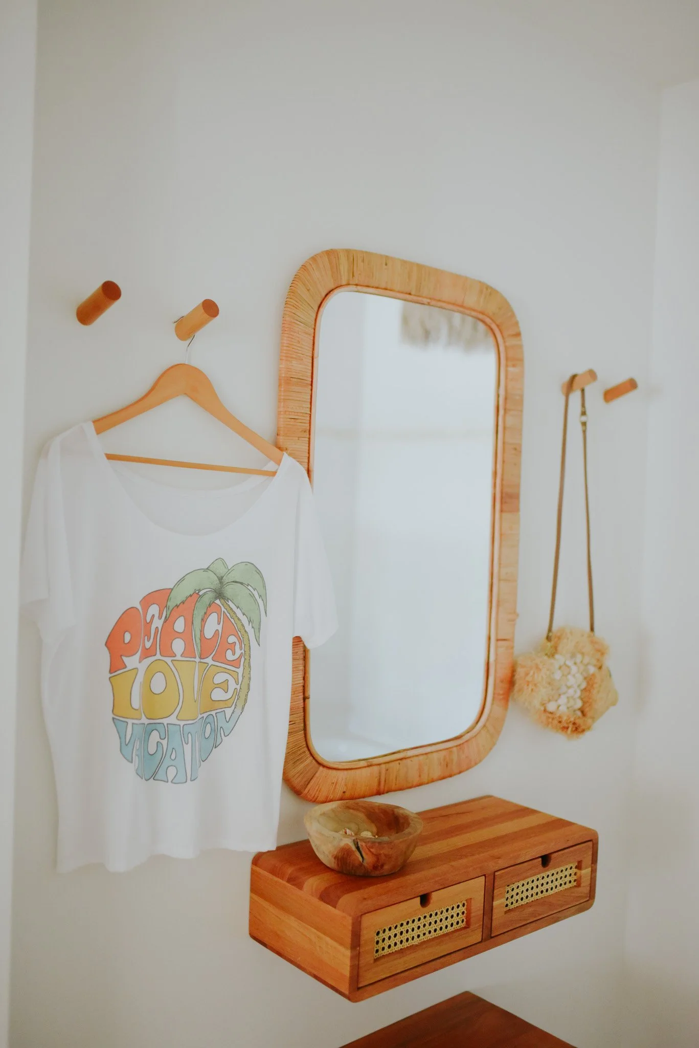 A white t-shirt with a colorful print hanging on a wooden hook next to a mirror, a wooden floating shelf with a small decorative bowl, and a fluffy bag hanging on another hook in a minimalist room with white walls.