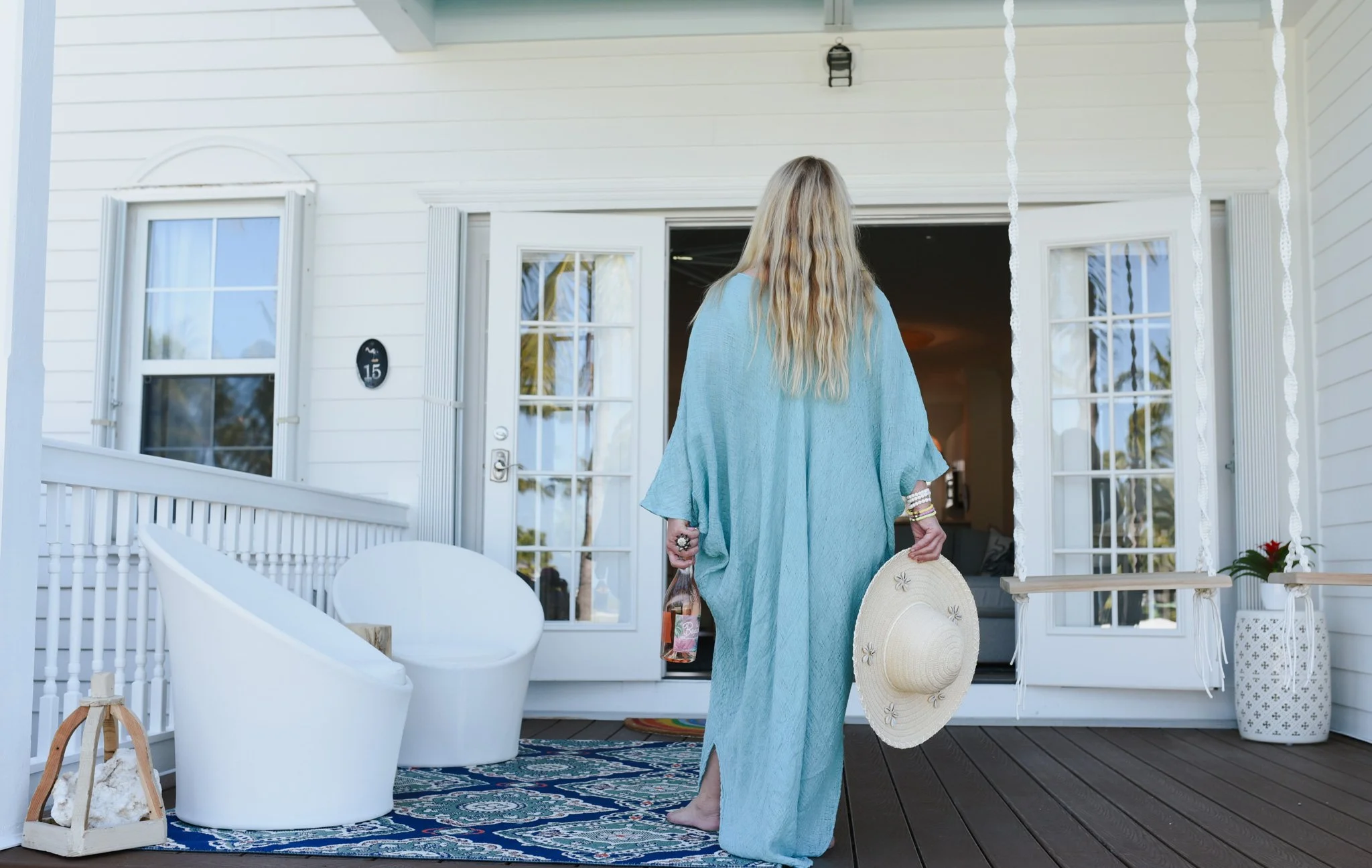 A woman in a blue cover-up holding a hat and a bottle of drink on a porch, facing an open door of a white house with glass panes.