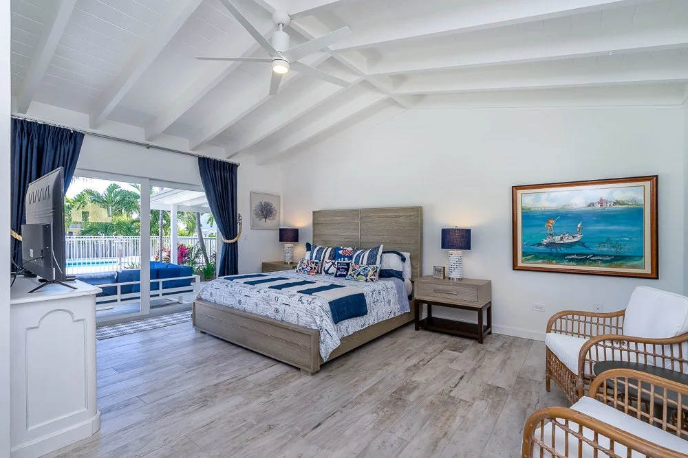 A beachfront bedroom with a large bed, nightstands with lamps, a framed painting of a boat on water, a white armchair, and a sliding glass door leading to a patio with a pool and tropical greenery.
