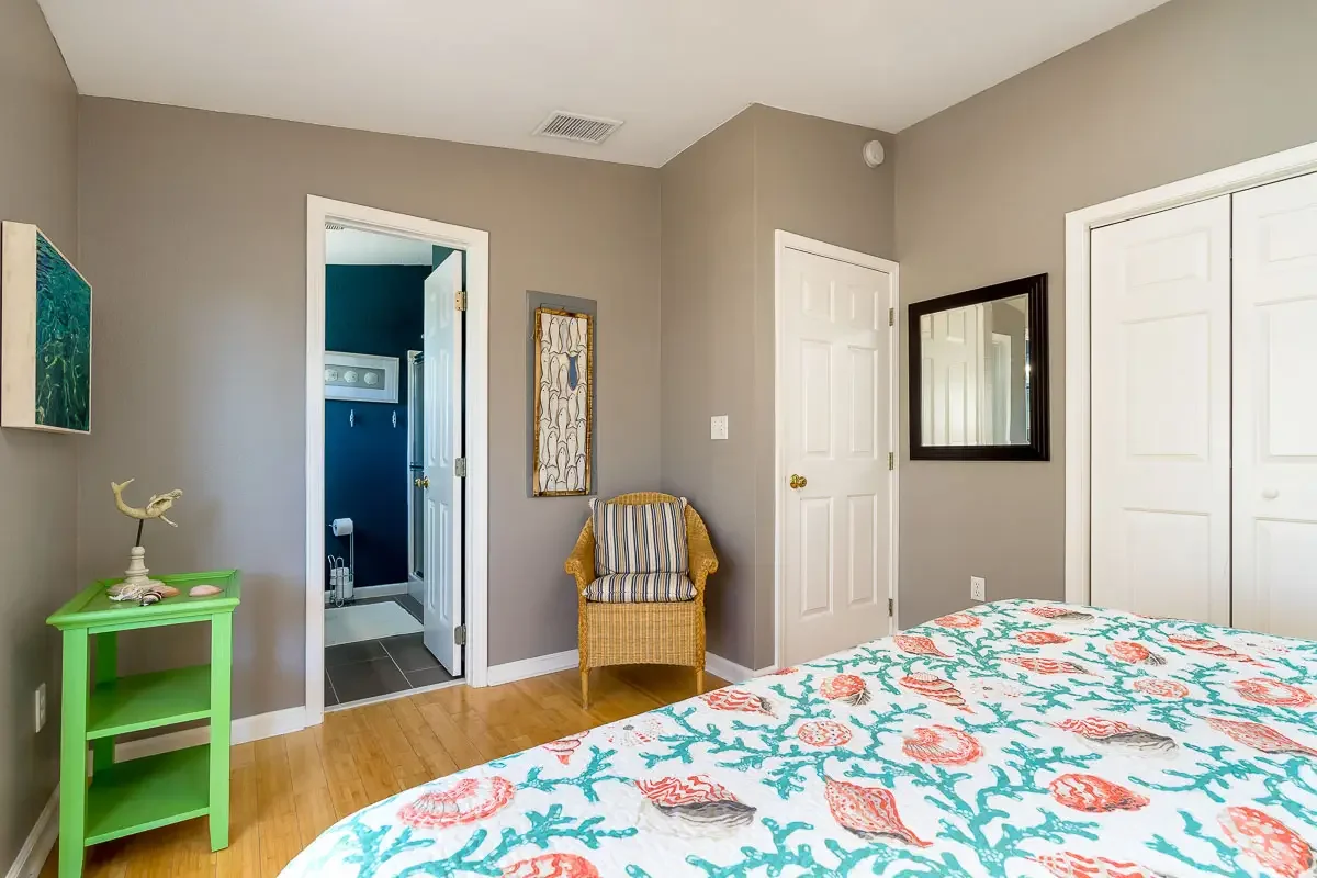 A bedroom with a bed covered in a seashell and coral patterned quilt, a yellow wicker chair with a striped cushion, a green side table with a decorative whale figure, and a wall mirror. There are two closed white closets and a door leading to an adjo