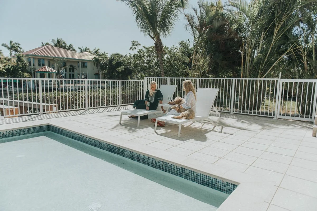 Two women sitting on poolside chairs near a swimming pool, with a dog on one woman's lap at a house with trees and palm trees in the background.