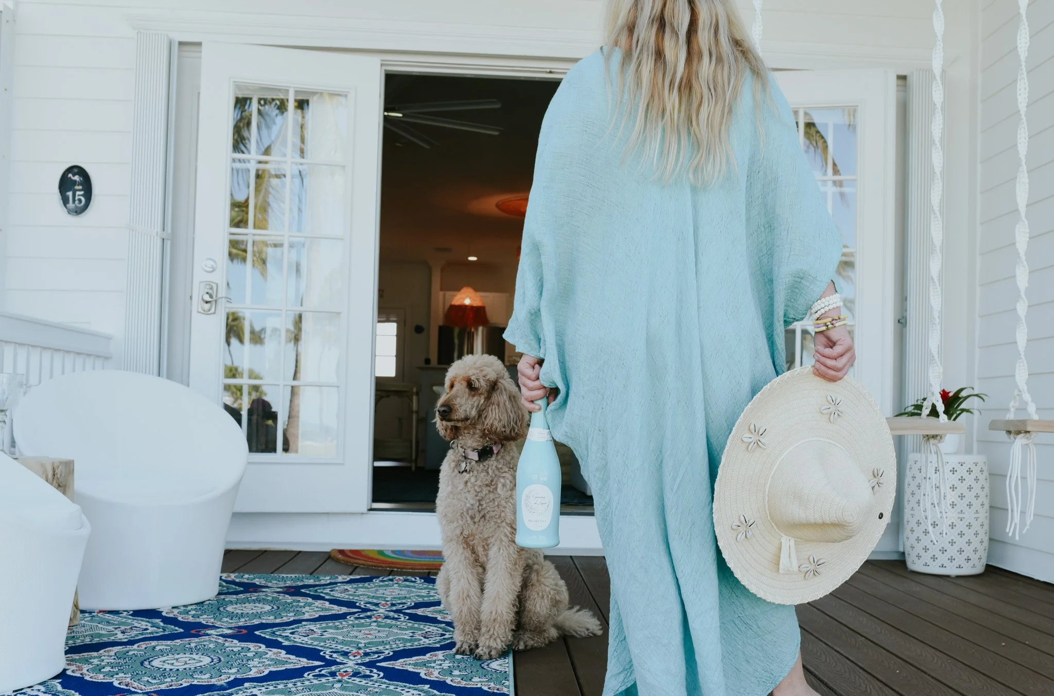 A woman in a light blue cover-up holding a straw hat and a bottle of sparkling wine, standing on a porch with a poodle sitting next to her. The porch has white furniture and a colorful rug, with an open door leading into the house.