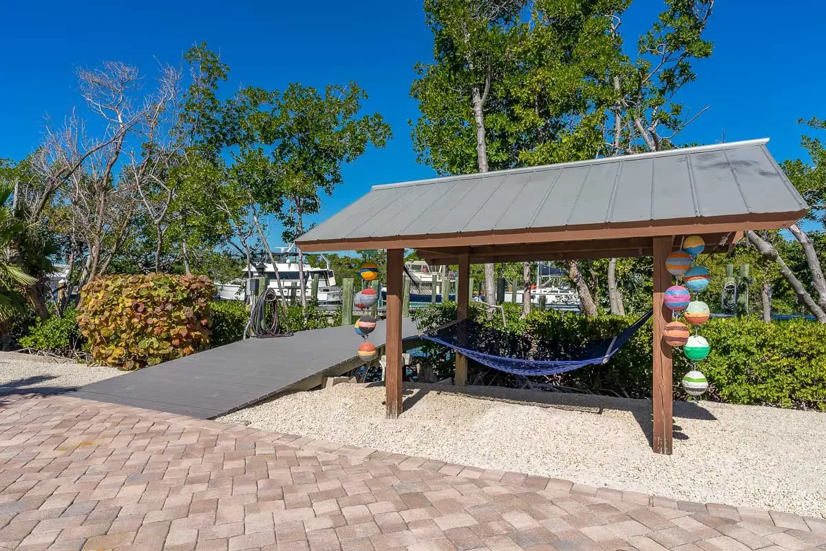 A small wooden shelter with a metal roof and a blue hammock underneath, decorated with colorful hanging lanterns, situated in a landscaped seaside area with boats in the background and clear blue sky overhead.