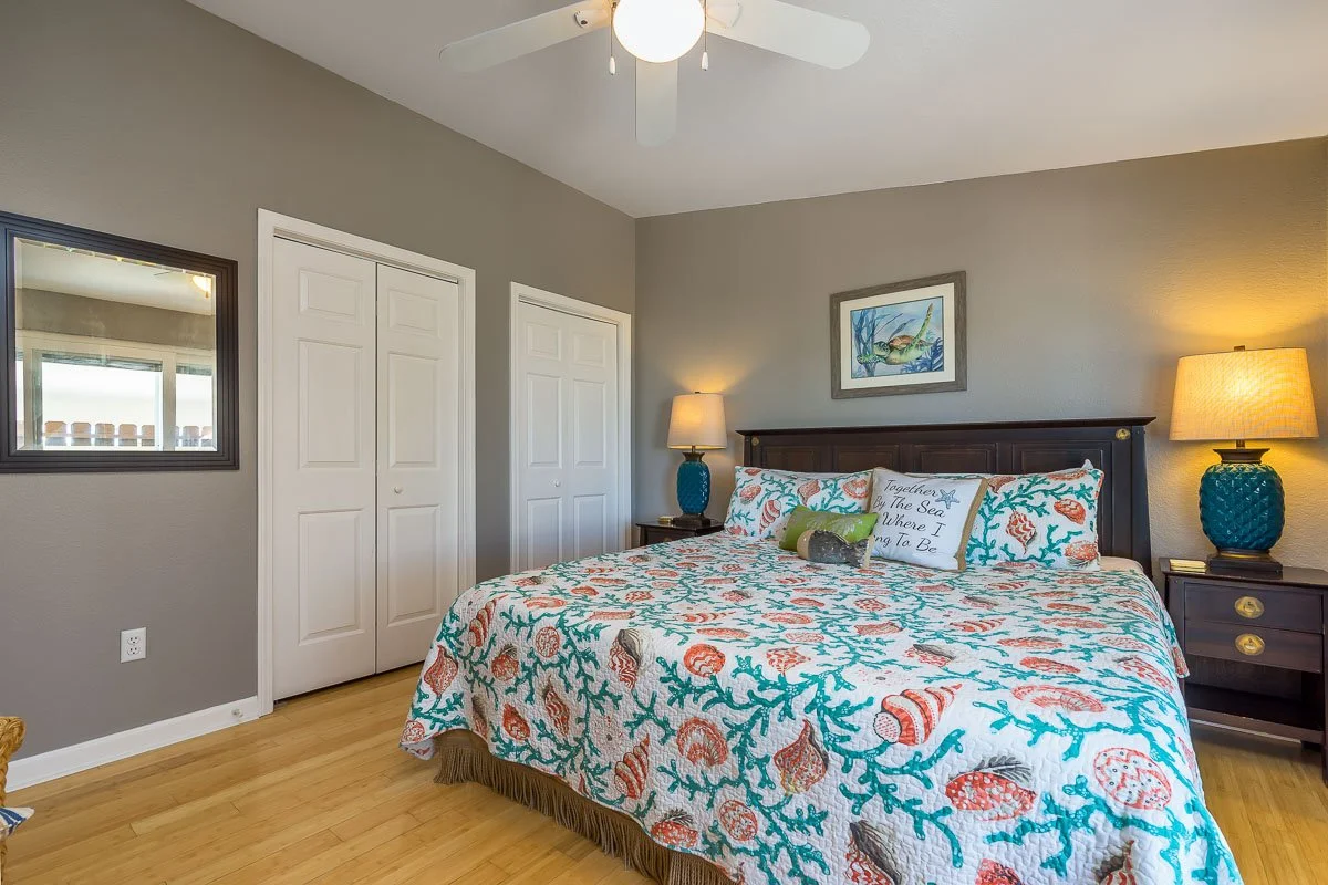 A bedroom with a bed covered in a shell and coral patterned quilt. There are two matching blue lamps on bedside tables, a framed fish picture above the bed, and a decorative pillow with sea-themed text. A ceiling fan hangs from the white ceiling, and