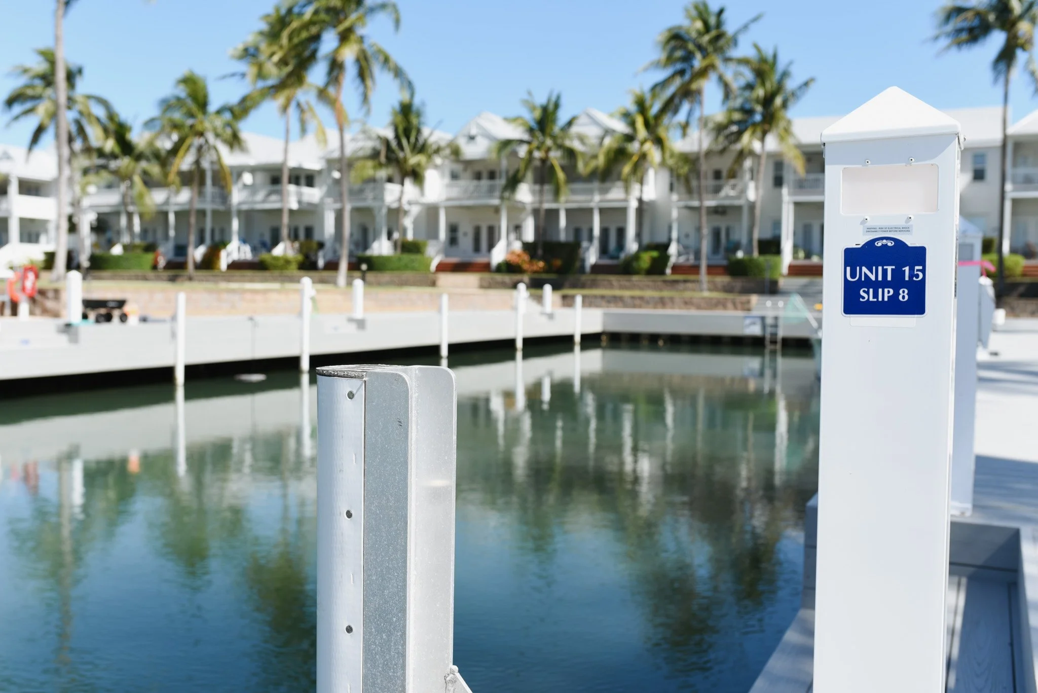A marina with a dock, water, and white buildings with balconies and palm trees in the background.
