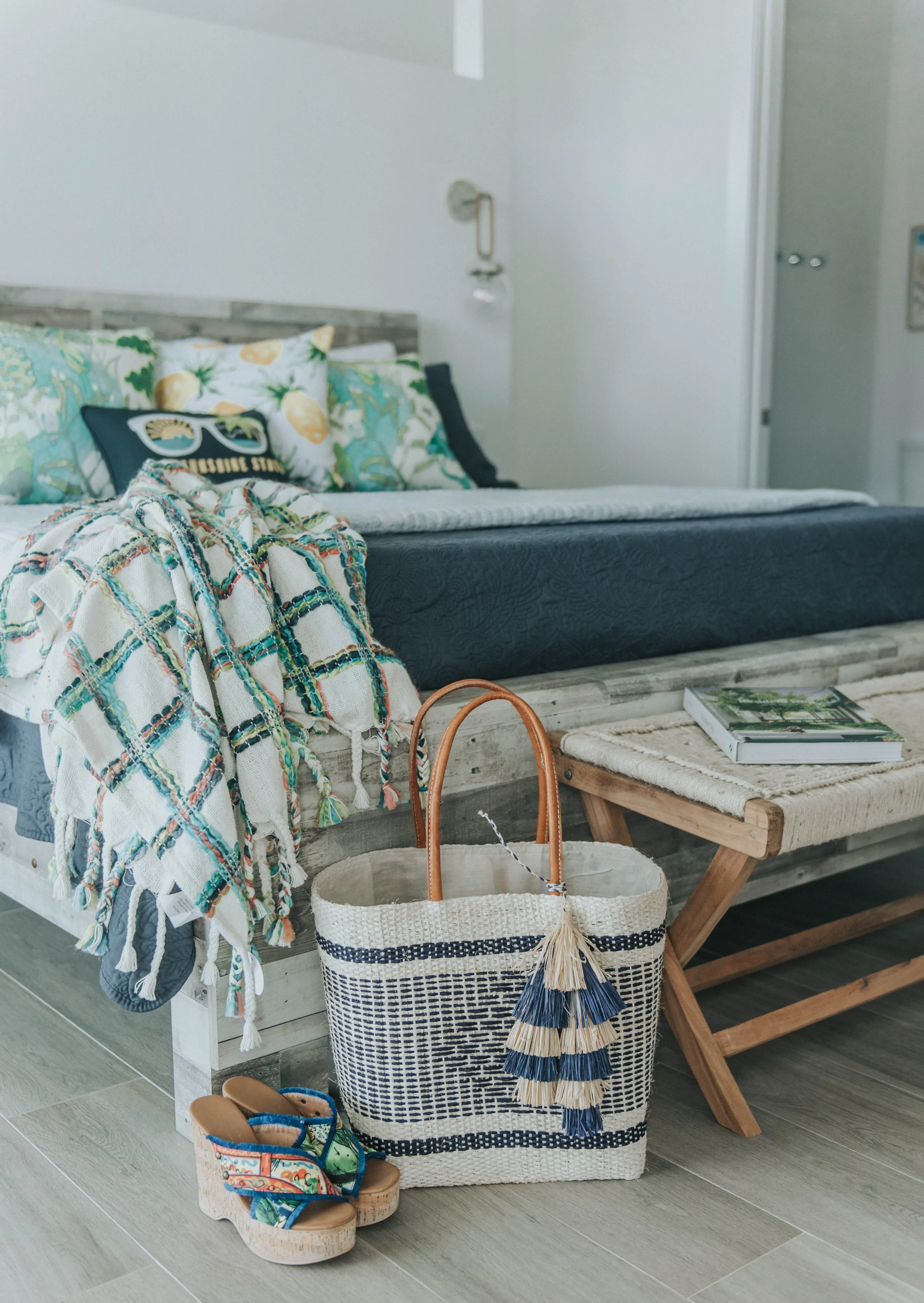 Coastal bedroom with a bed that has colorful pillows and a dark blue bedspread, a blanket with tassels draped over the side, a woven tote bag with a tassel on the floor, a pair of colorful wedge shoes, a small wooden bench with a book on top, in fron