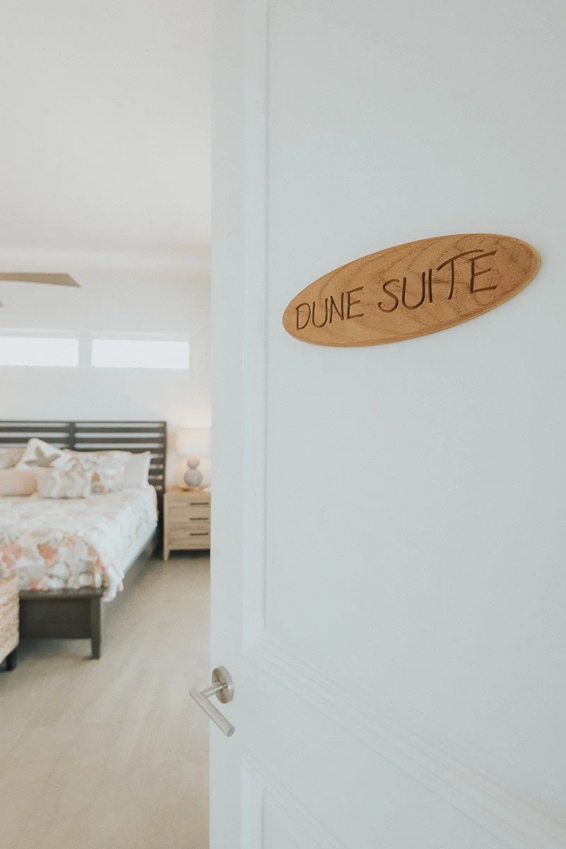 A bedroom seen through an open door with a wood sign reading 'DUNE SUITE' on the wall.