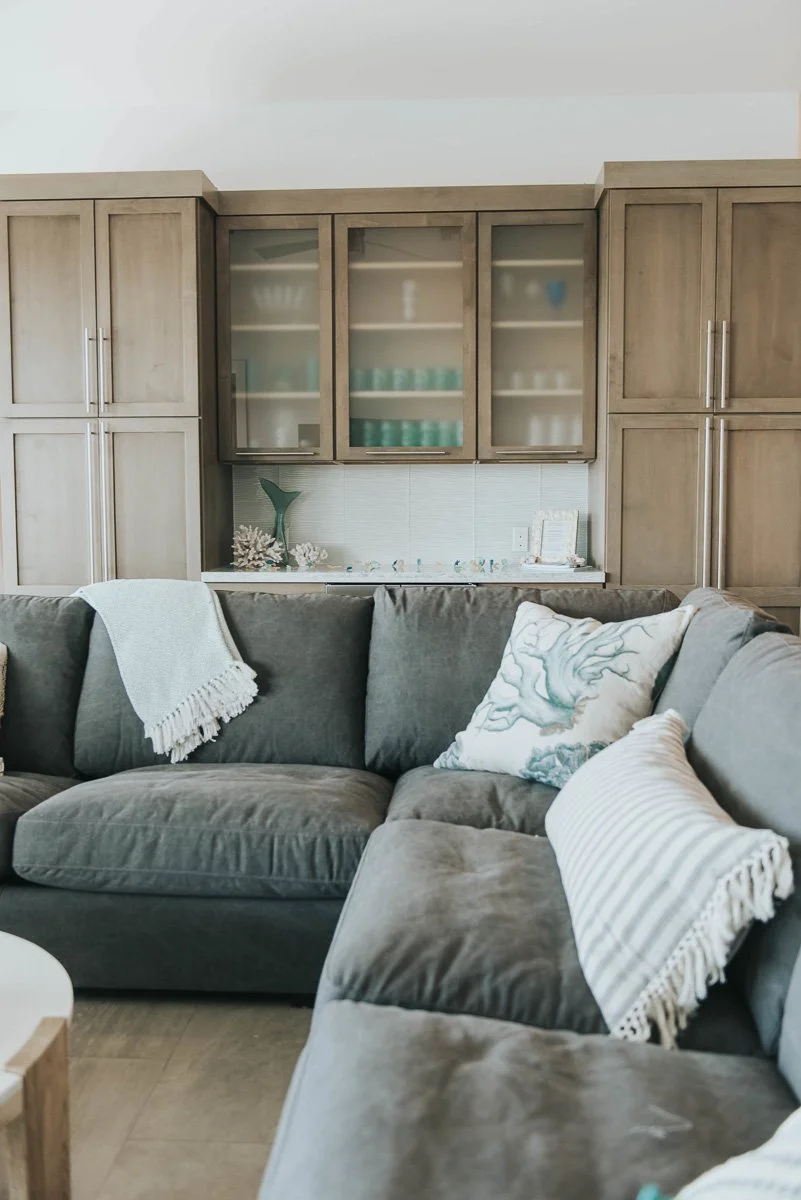 Living room with a gray sectional sofa decorated with white pillows and throws, and wooden cabinets with glass doors in the background.
