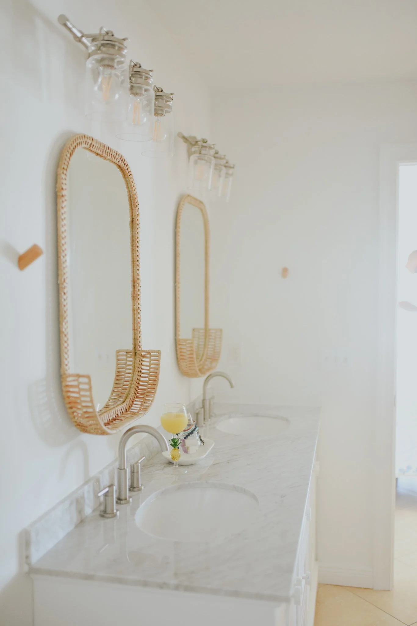 A bathroom vanity with a marble countertop, two sinks, and two wall-mounted wicker mirrors. Above each mirror are glass light fixtures. On the countertop, there is a small tray holding a glass of yellow juice with a decorative pineapple and a small f