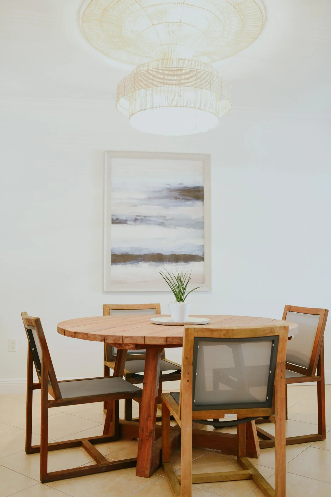 A round wooden dining table with four wooden chairs, one of which has a black mesh back, in a room with white walls. A potted plant is on the table, and a large abstract painting hangs on the wall behind it. A hanging light fixture is above the table