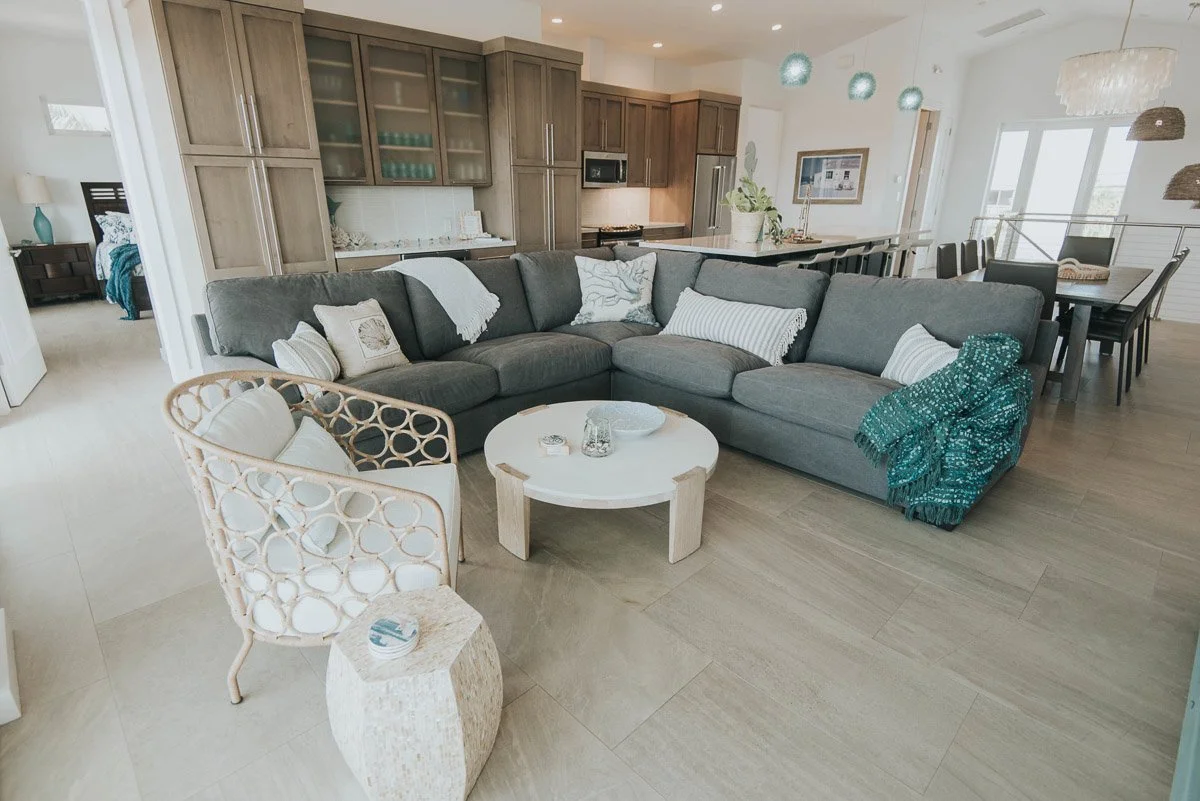 Open-concept living room with a gray sectional sofa, white pillows, a round white coffee table, a beige woven armchair, and a small beige side table. The background shows a kitchen with wooden cabinets and a dining area with a black table and chairs,