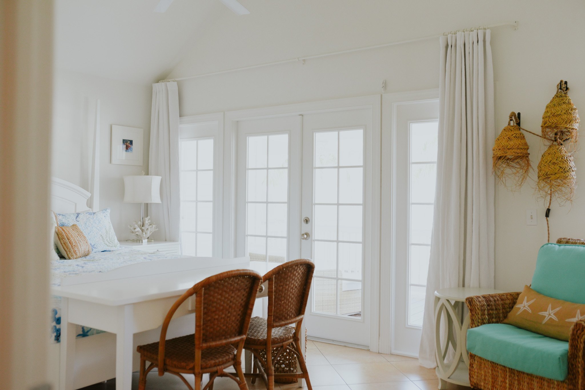 Bright tropical hotel room with white walls and large glass doors leading outside, decorated with nautical and beach-themed furniture and accessories.