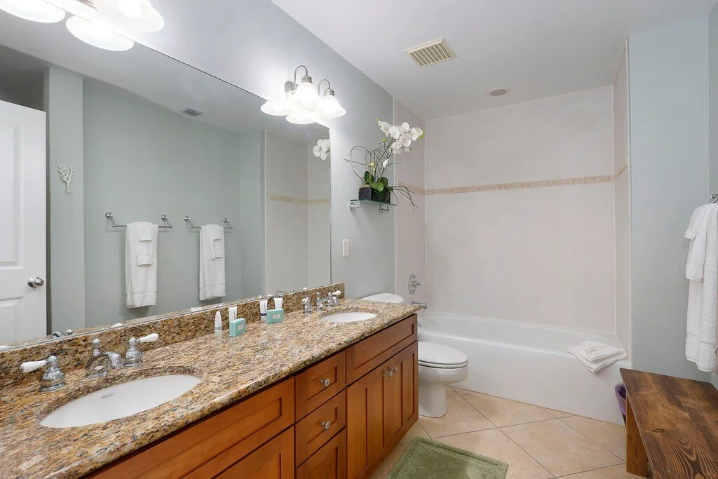 Bathroom with double sink granite countertop, large mirror, white towel racks, bathtub with shower, and a small wooden bench. Light fixtures above mirror, and plants near the ceiling.