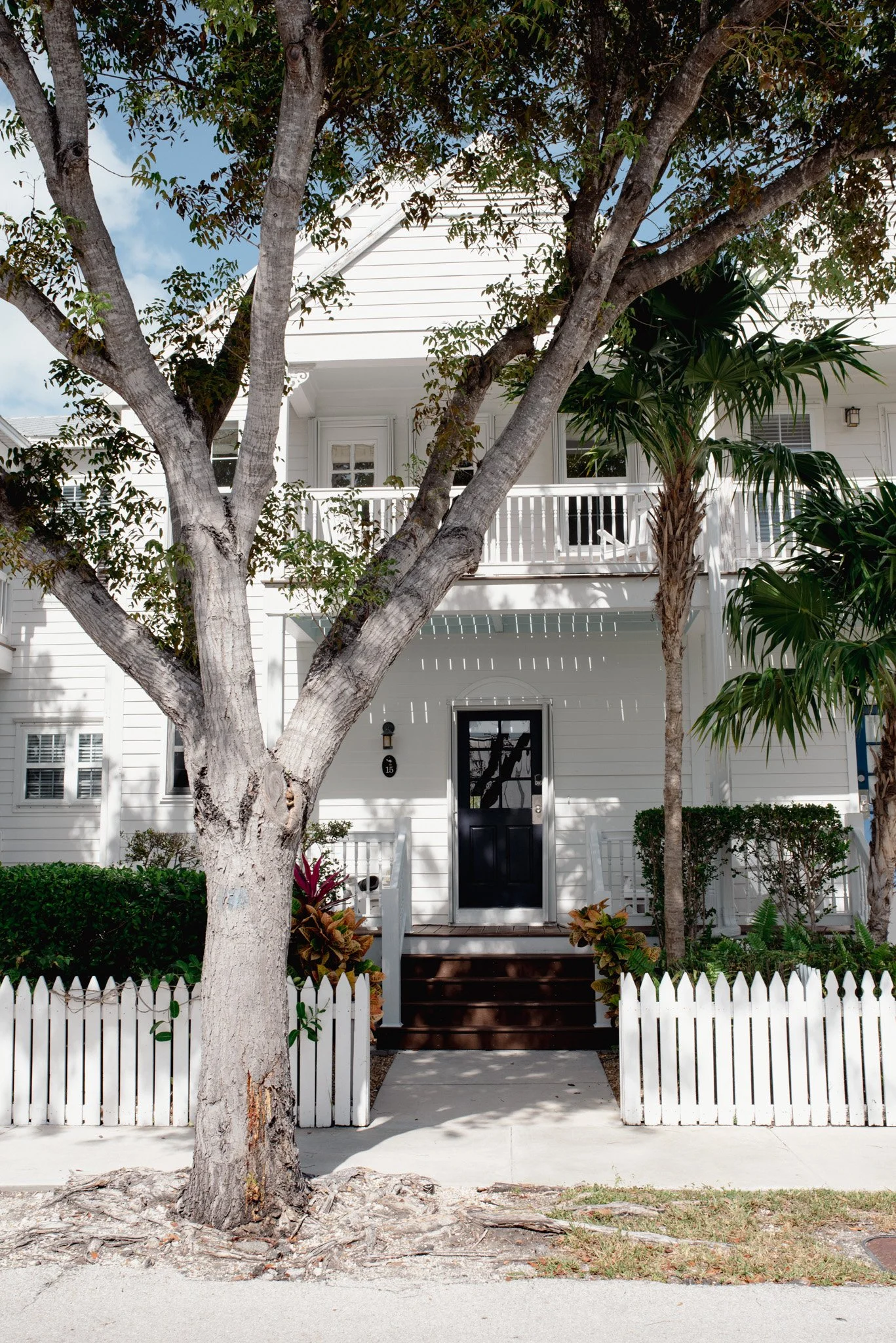 Front view of a white two-story house with a black door and a small front yard, trees, and a white picket fence.