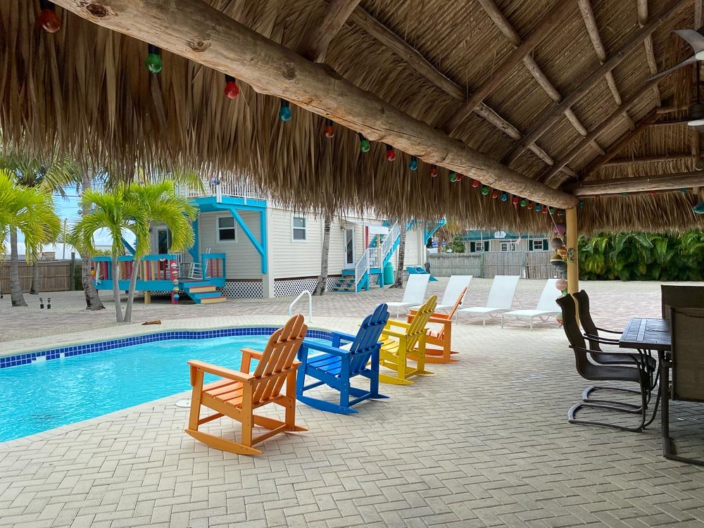 A backyard pool area with a thatched roof shaded sitting area, colorful Adirondack chairs, a swim-up bar, and a small blue house with deck, surrounded by palm trees and a wooden fence.