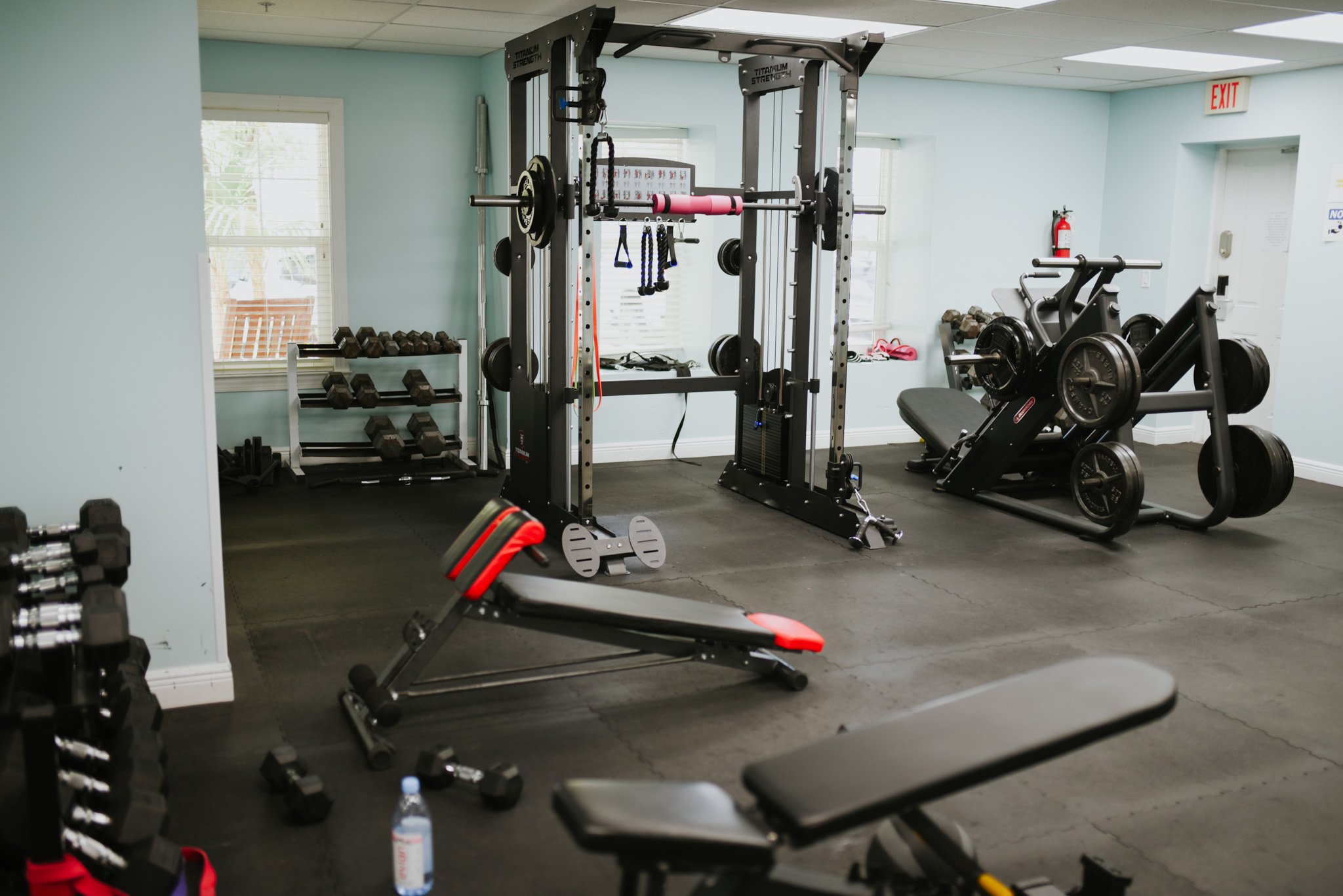 A gym with various workout equipment including dumbbells, a squat rack with barbells, a rowing machine, a weight bench, an adjustable workout bench, and a water bottle on the floor.