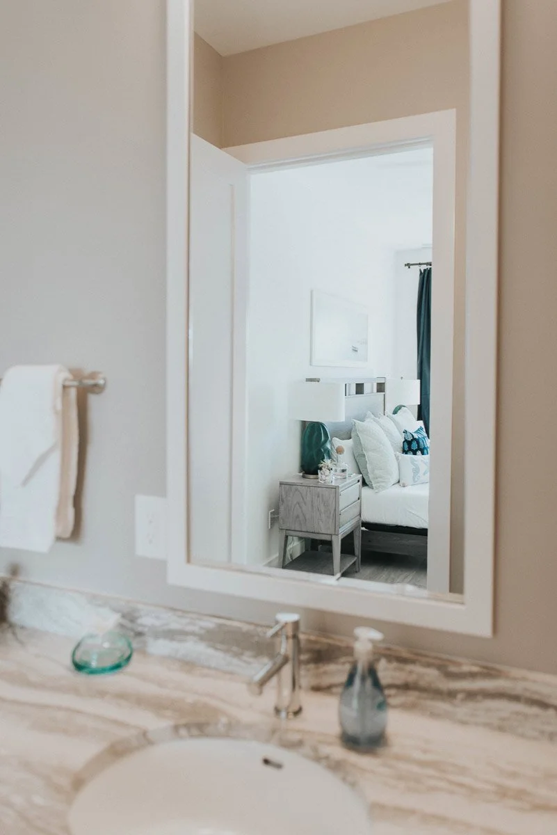 A bathroom sink with a beige marble countertop and a mirror reflecting a bedroom with a bed, nightstand, and lamps.