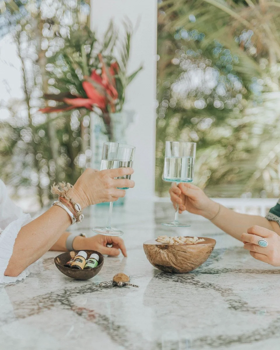 Two people holding glasses of water toasting at a marble table with bowls of nuts and essential oils, with greenery in the background.