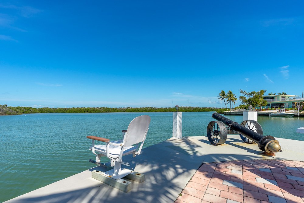 A cannon on a pier overlooking a waterway with palm trees and houses in the background.