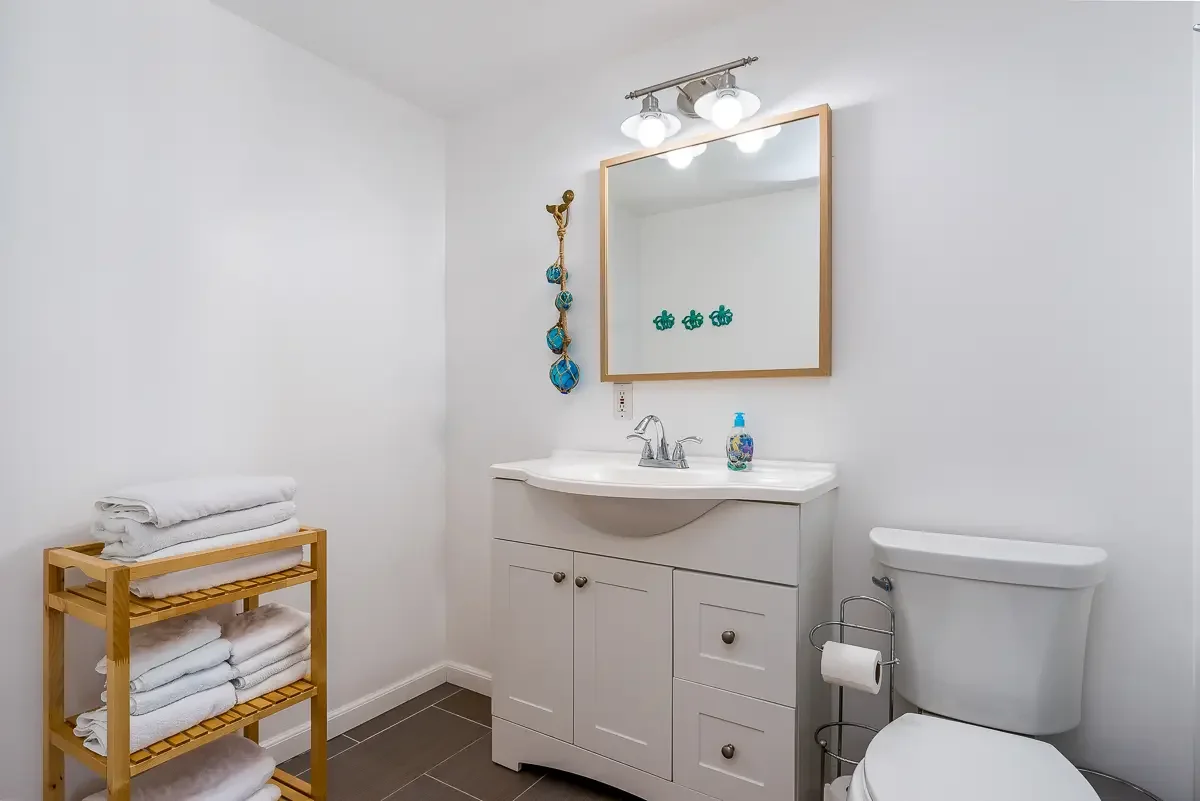 A clean, white bathroom featuring a vanity with a mirror, three bulb light fixture, a small bottle of hand soap, a toilet, and a towel rack with three shelves holding rolled white towels. The walls are white, and the floor has dark tiles.