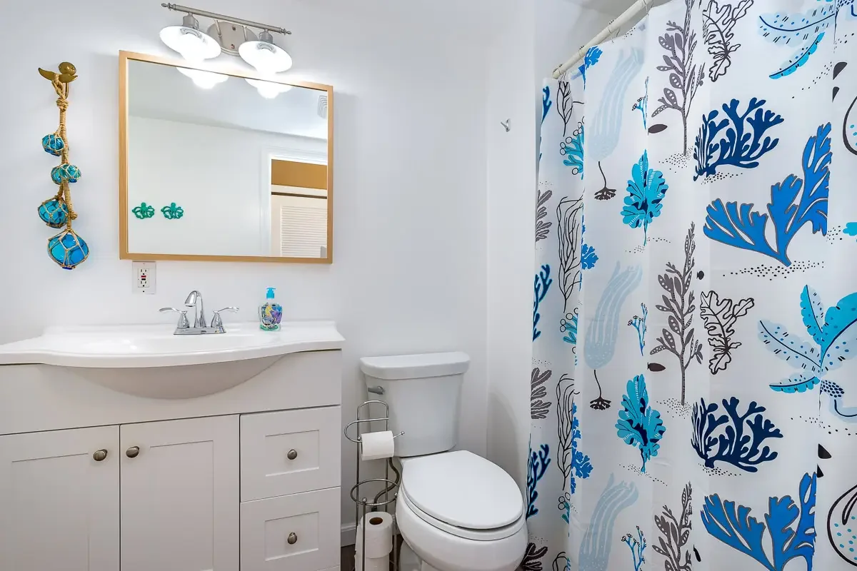 A small bathroom with a white vanity, a mirror, a toilet, a shower with a blue and white patterned curtain, and decorative wall hooks shaped like fish.