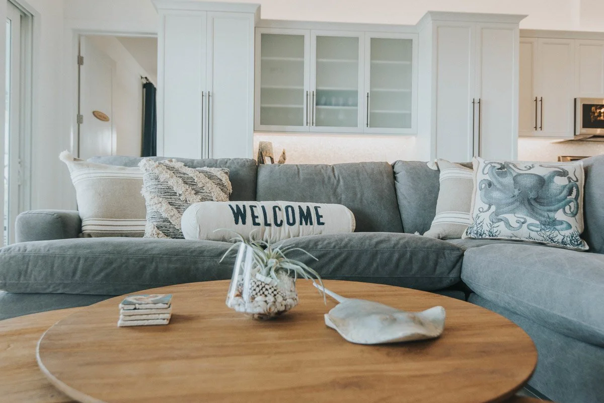 Living room with a gray sectional sofa, decorative pillows, a wooden coffee table with a small plant and shells, and a background of white kitchen cabinets.