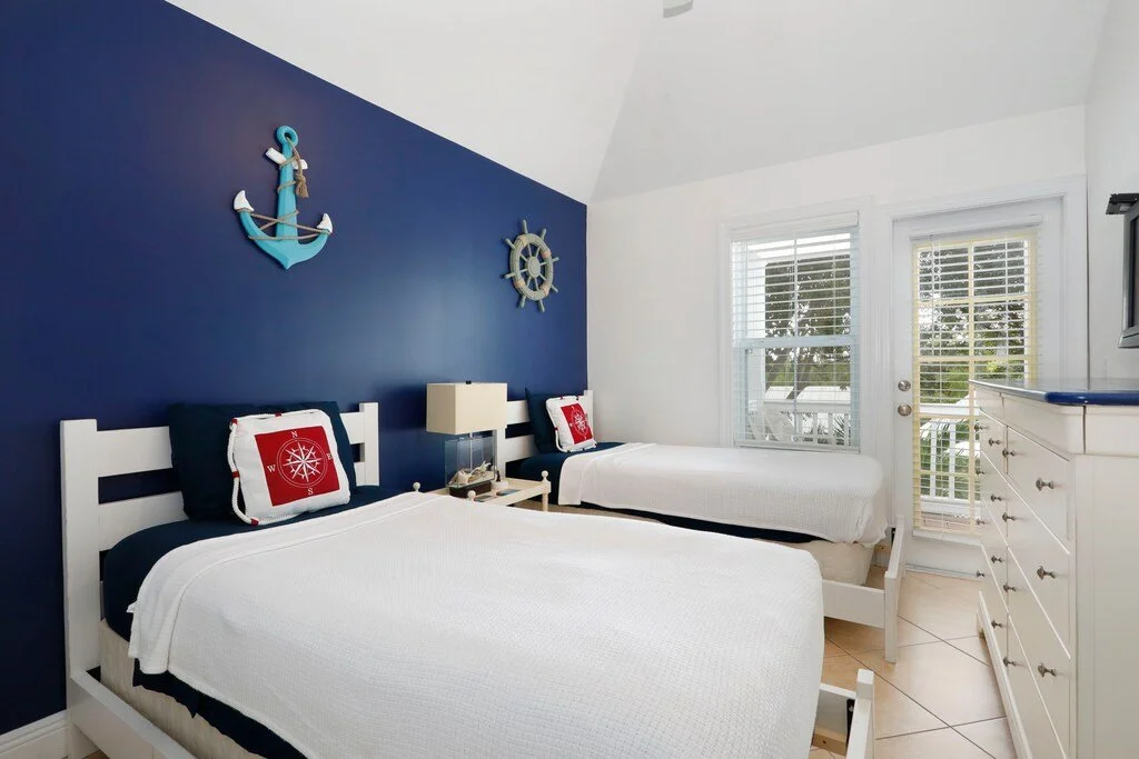 Beach-themed bedroom with two twin beds, navy and white decor, nautical wall art, and a door leading to a balcony.
