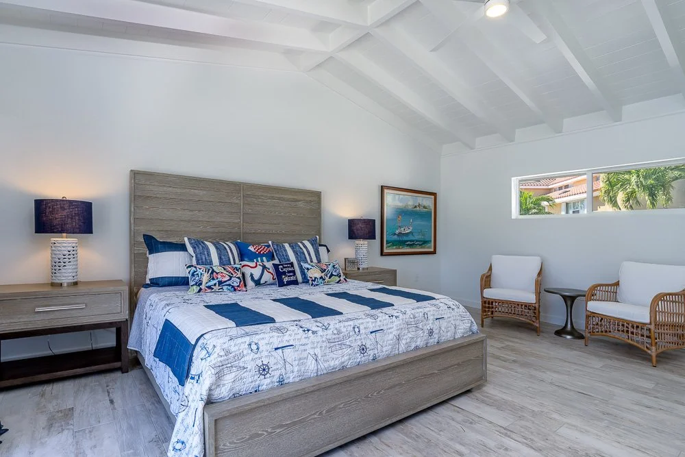 A spacious bedroom with a large bed, nautical-themed bedding, two nightstands with lamps, a framed ocean painting, two white armchairs, a small dark side table, a horizontal window showing palm trees outside, and a white vaulted ceiling.