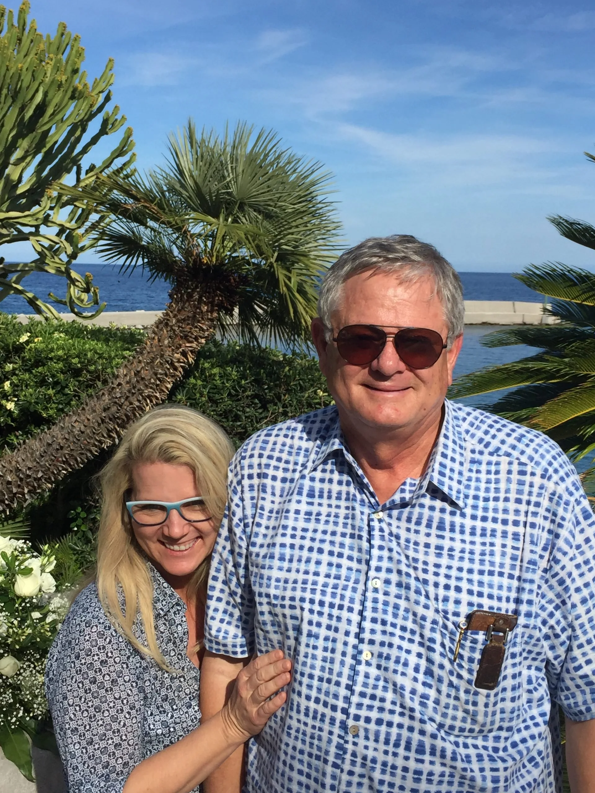 A smiling man and woman pose outdoors near the ocean, with palm trees, lush greenery, and a clear blue sky in the background.