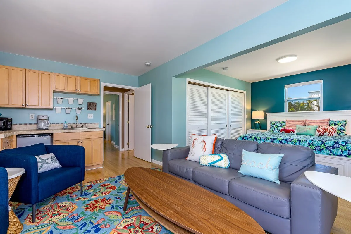 Coastal-themed living room and kitchenette in a vacation rental or apartment with blue walls, colorful rugs, a sofa with beach-themed pillows, a bed with tropical bedding, and wooden cabinets.
