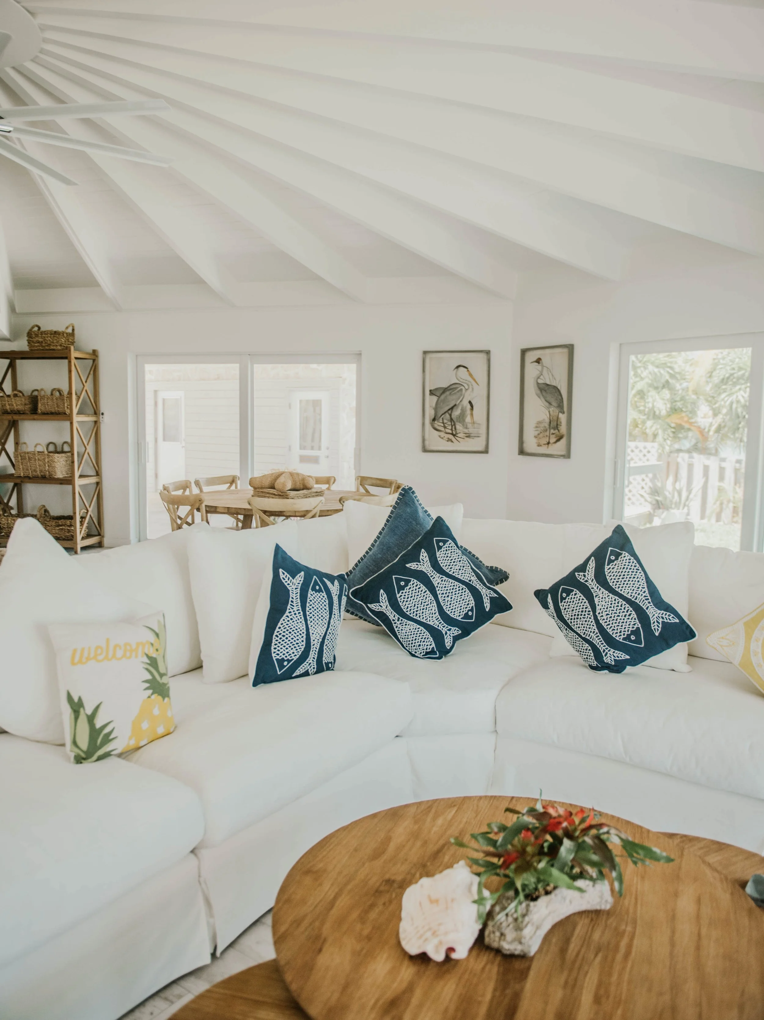 Bright living room with white sectional sofa decorated with blue and white fish themed pillows, a round wooden coffee table with shell and plant decoration, and a dining area with wooden table and chairs in the background.