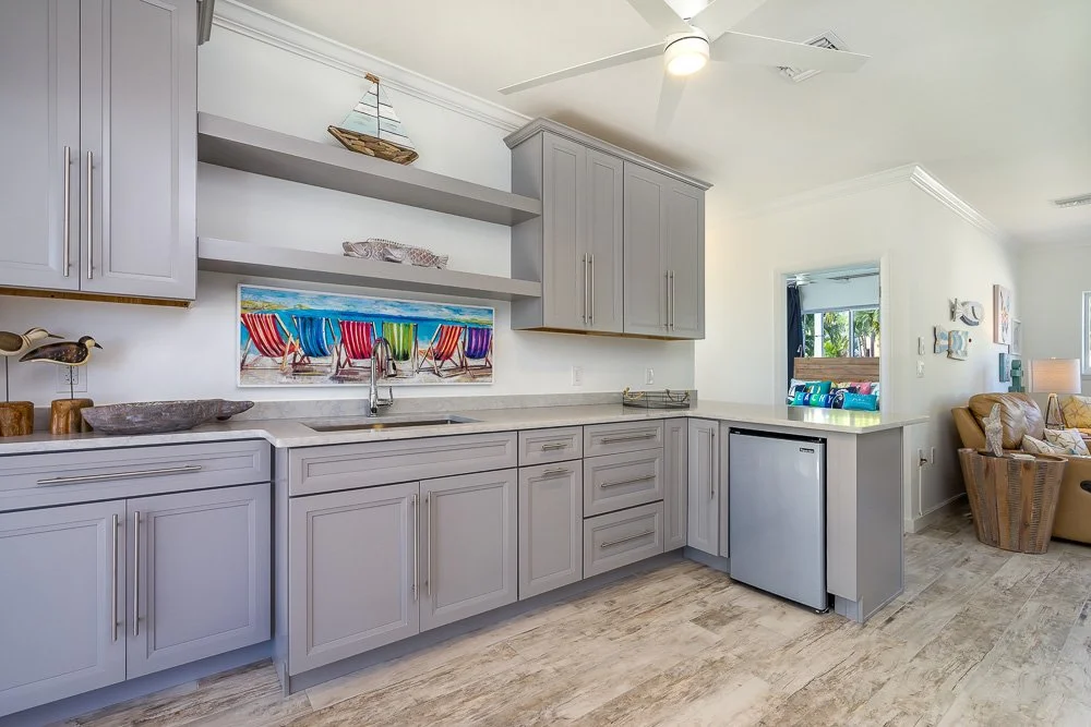 Kitchen area with light gray cabinets, open shelves with decorative items, colorful beach chairs painting, small refrigerator, and living room with beige sofa and wall art in background.
