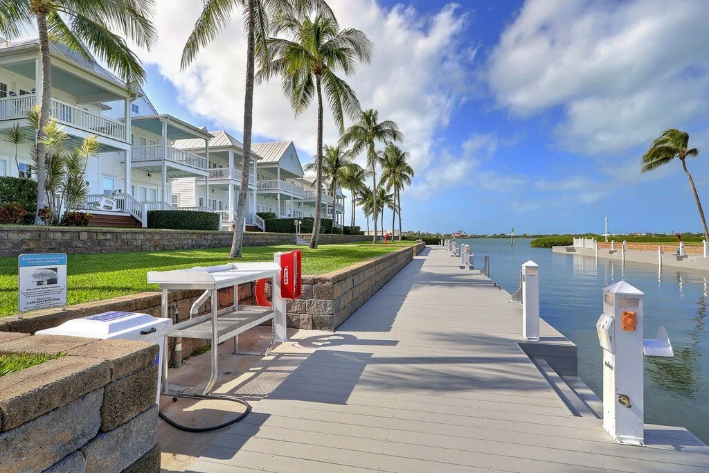 A wooden dock extends into a waterway beneath a partly cloudy blue sky. There are white houses with balconies and palm trees along the walkway, which has utility and safety equipment.