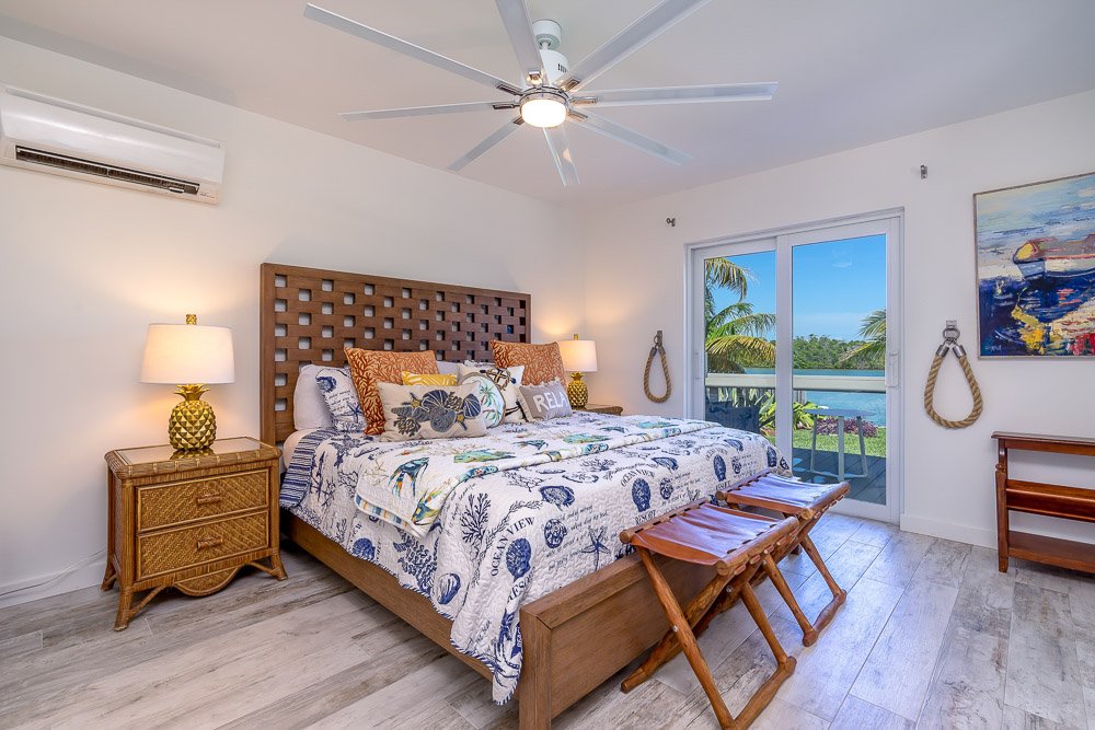 A bedroom with a large bed featuring ocean-themed bedding, wooden furniture, and a view of palm trees through sliding glass doors leading to a balcony.