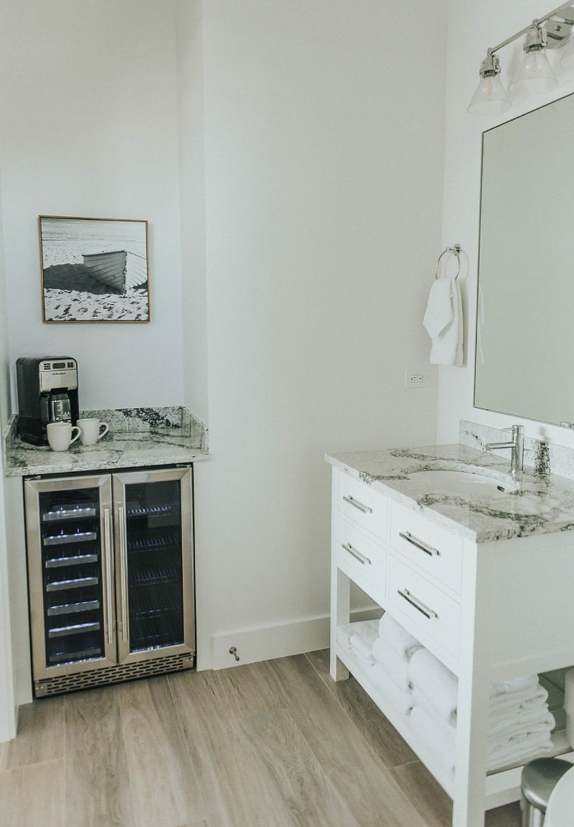 A modern bathroom or wet bar area with white walls, a marble countertop, a small wine cooler, a coffee machine, a framed black and white beach scene artwork, a white towel on a ring, a large mirror, and a light fixture with glass shades.