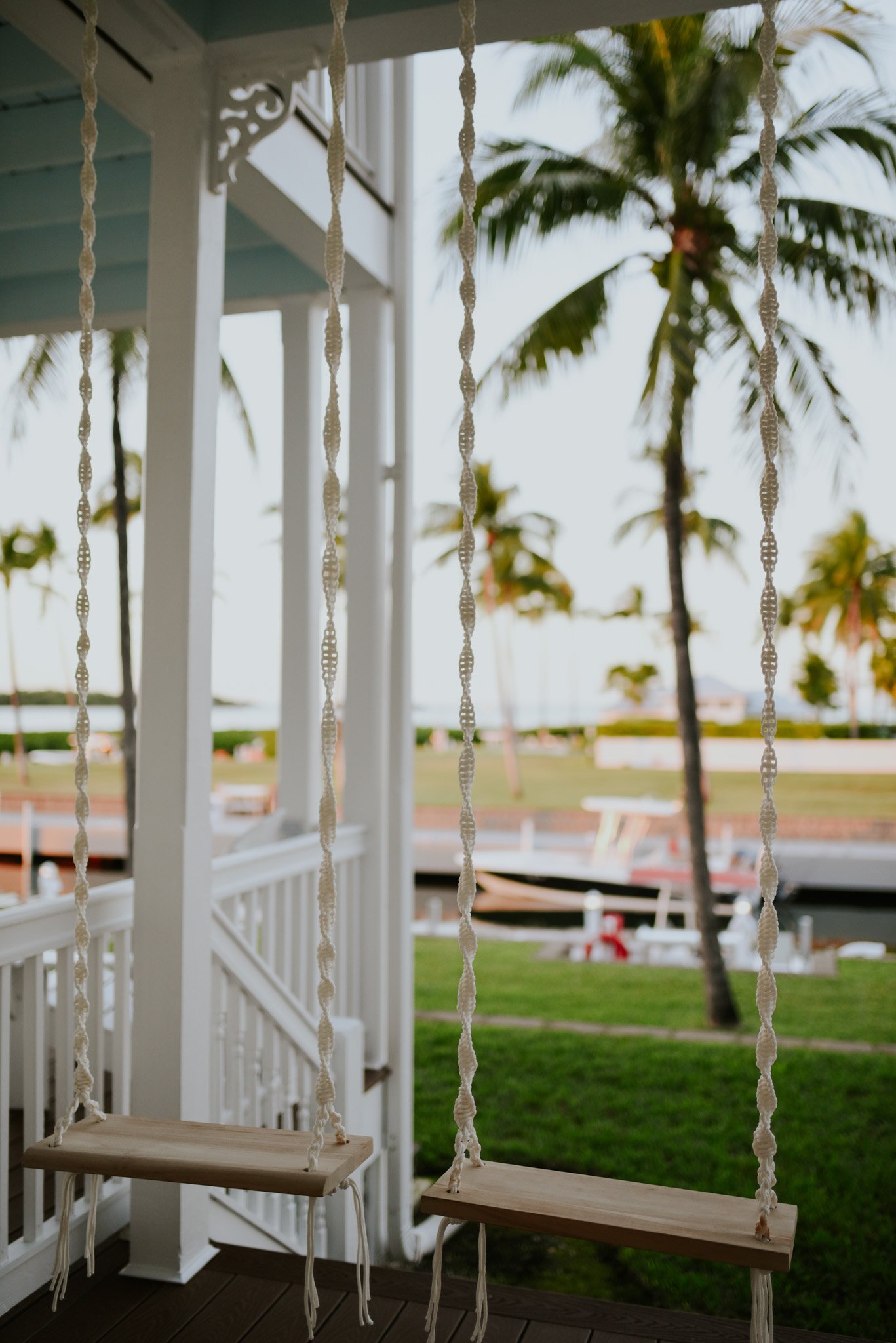 Two wooden swings hanging from a porch ceiling with white ropes, overlooking a tropical outdoor scene with palm trees, grass, and a walkway or river in the background.