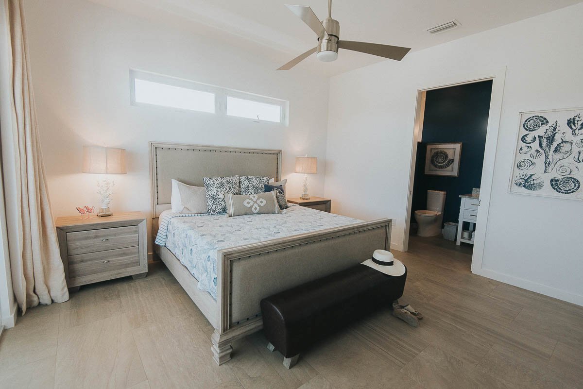 A modern bedroom with a large bed, beige upholstered headboard, and matching side tables with lamps. There is a black bench at the foot of the bed, topped with a hat. The room has light wooden flooring, a ceiling fan, and a window above the bed.