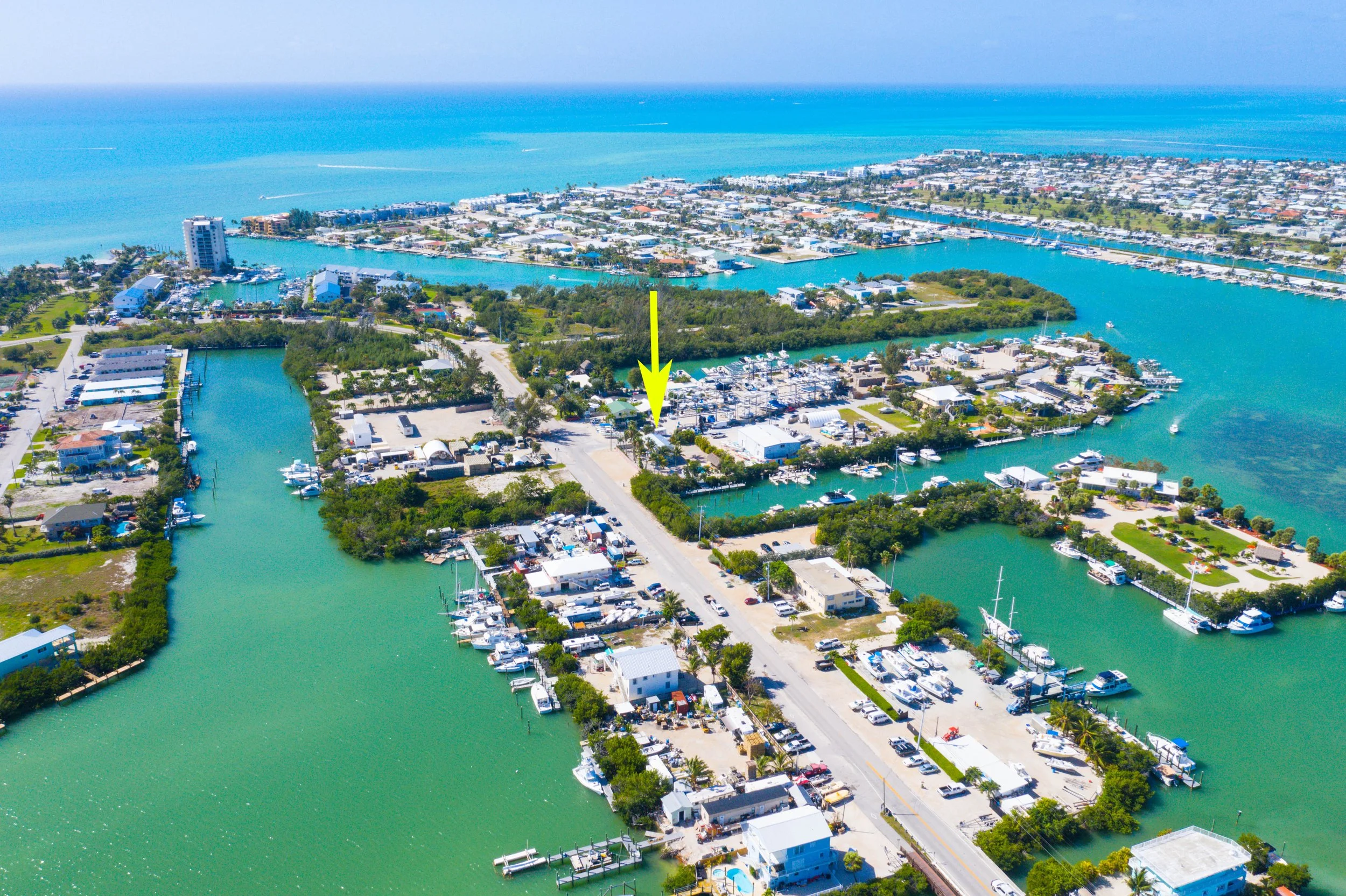 Aerial view of a marina with boats, water channels, and buildings, with the ocean in the background. An arrow points to a specific location on the marina.