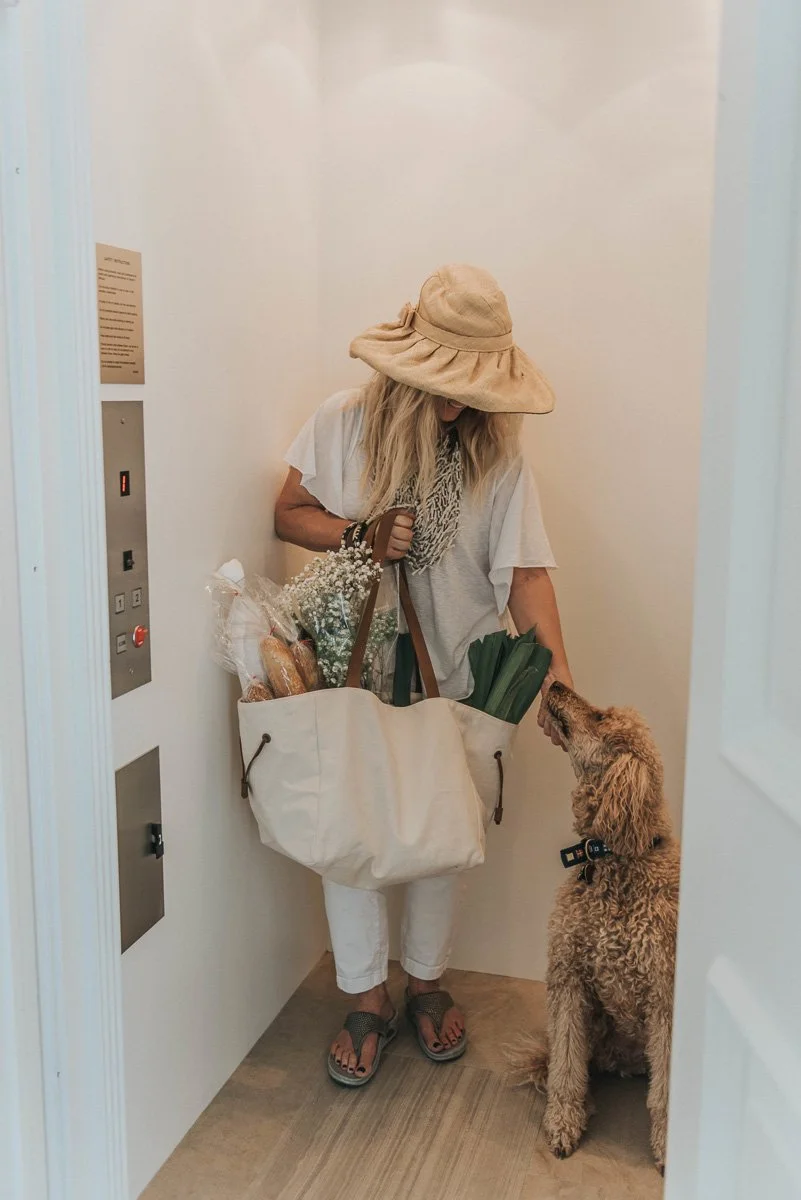 A woman with long blonde hair wearing a large floppy hat, white shirt, and white pants stands in an elevator, holding a large tote bag filled with groceries including bread, flowers, and leafy greens. She is petting a curly-haired dog, likely a poodl