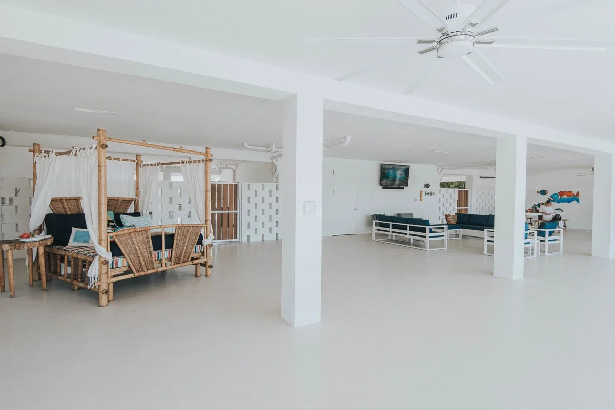 Spacious indoor lounge area with tropical decor, including wicker furniture and a four-poster bed with white curtains, white walls, and minimal decor.